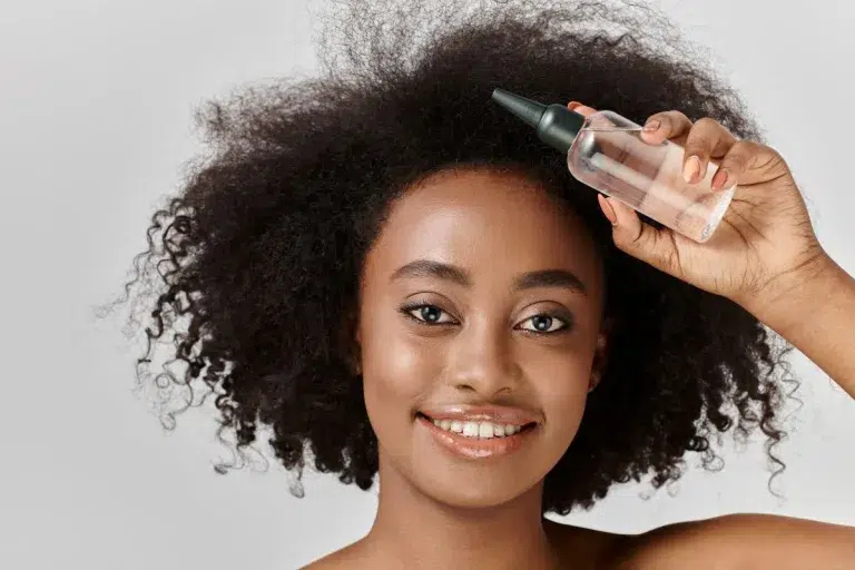 Gros plan d'une femme souriante appliquant une huile capillaire sur son cuir chevelu.