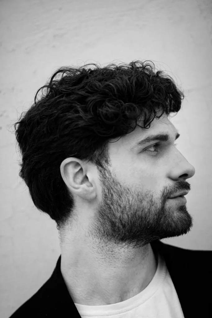 Homme de profil aux cheveux mi-longs ondulés coiffés vers l'arrière, barbe courte